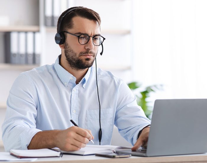 Serious mature guy in glasses and headphones works at computer, support clients, watches webinar or online lesson